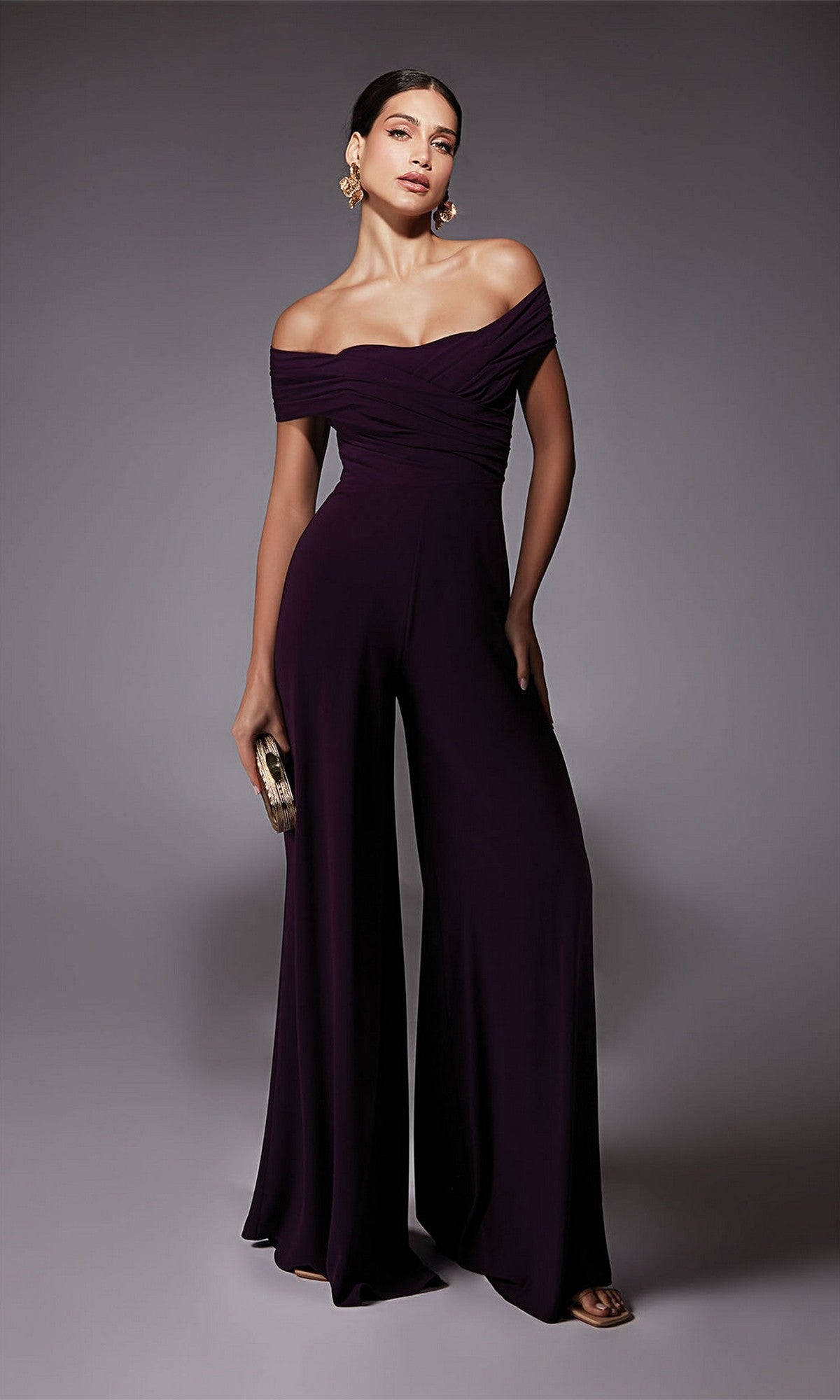 A woman wearing the Alyce 70067 long formal dress in dark purple poses against a plain background, holding a small clutch and gold earrings.